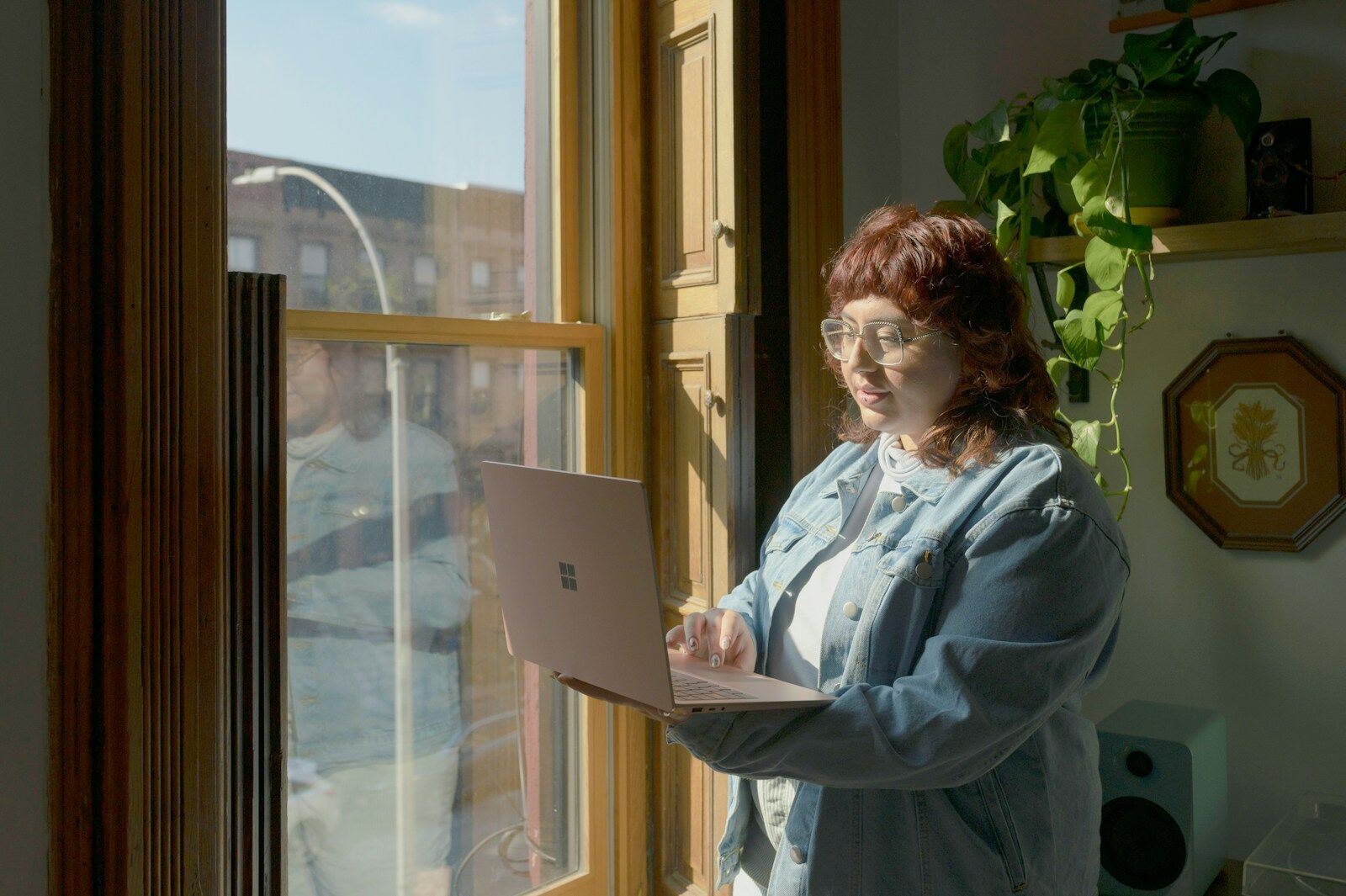 A person standing by a sunny window, working on a laptop held in one arm.