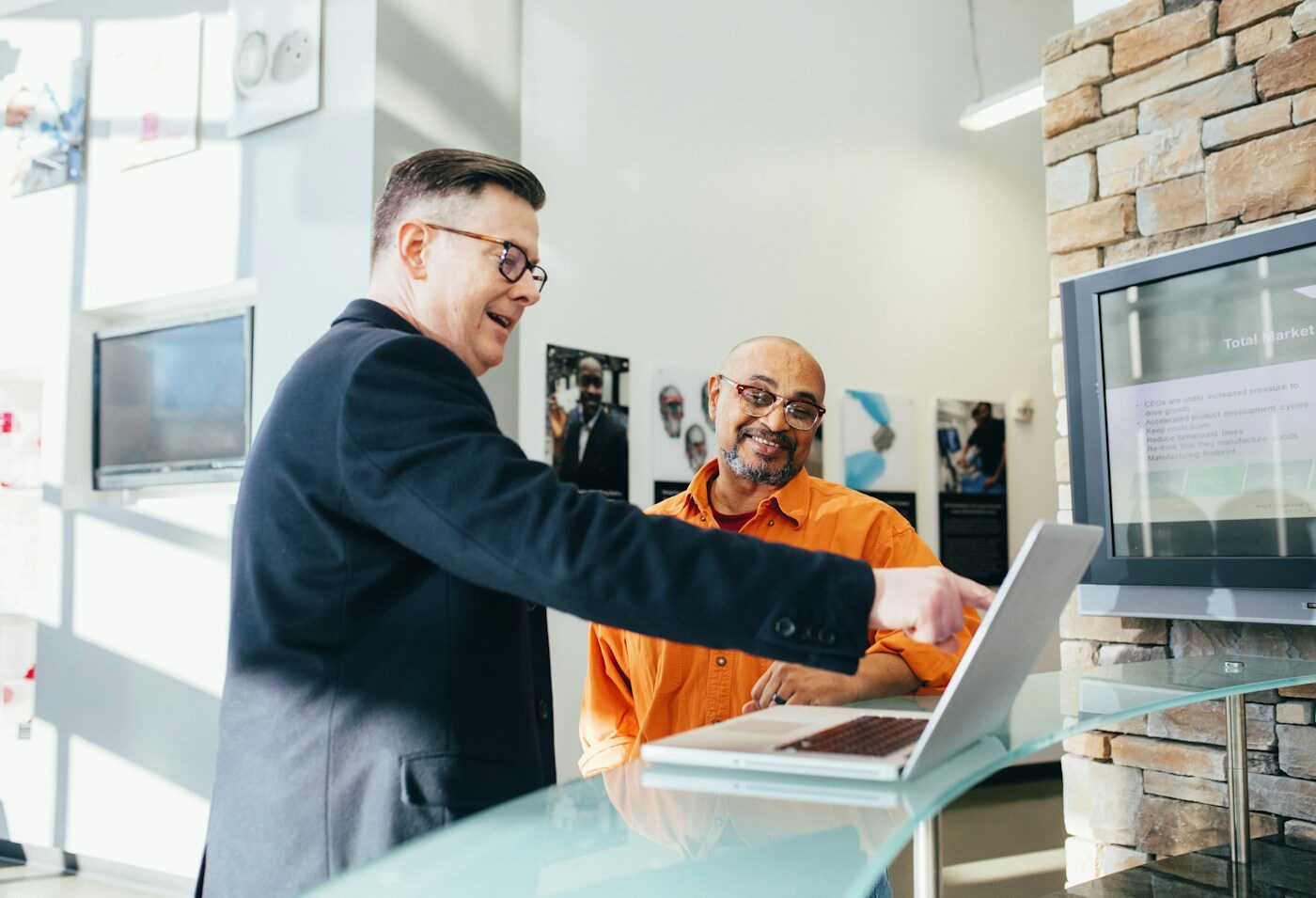 Two salespeople at a laptop in a bright office, one pointing at the screen while the other listens.